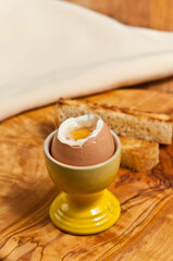 top, front view of a soft boiled egg with end cut off, exposing egg yolk and white, in a yellow egg dish with toast on rare wood plater 