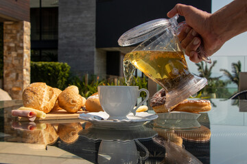 man serving tea during the day in white porcelain cup with bread and chocolate donuts in the background on a glass table in the background palm trees and clear sky