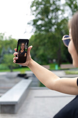 A white-skinned young girl in modern clothes is holding a smartphone and talking over a video link in a park in the summer.