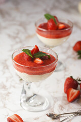 Several glasses with traditional creamy pudding dessert panna cotta with peppermint and strawberry jam surrounded fresh strawberries on a marble surface