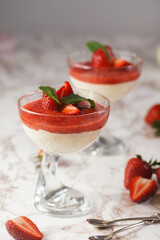 Several glasses with traditional creamy pudding dessert panna cotta with peppermint and strawberry jam surrounded fresh strawberries on a marble surface