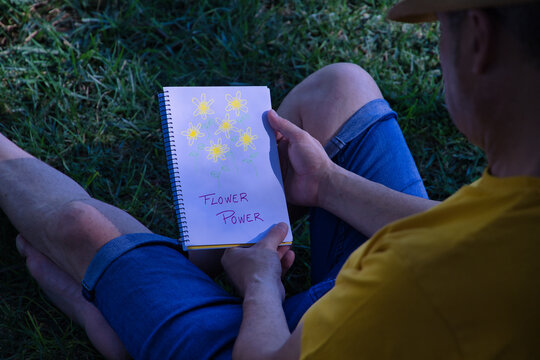 Hispanic mature adult man sitting on the lawn of a park and leaning on a tree with a notebook in his hands where he has drawn the motto the power of flowers.