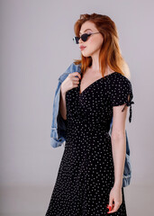 A dark-haired girl in a black dress stands on a white background with a denim jacket in her hands.