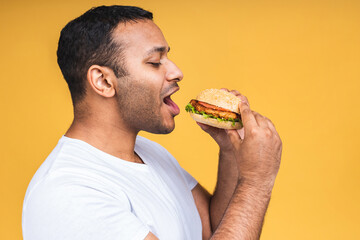 Young african american indian black man eating hamburger isolated over yellow background. Diet concept.