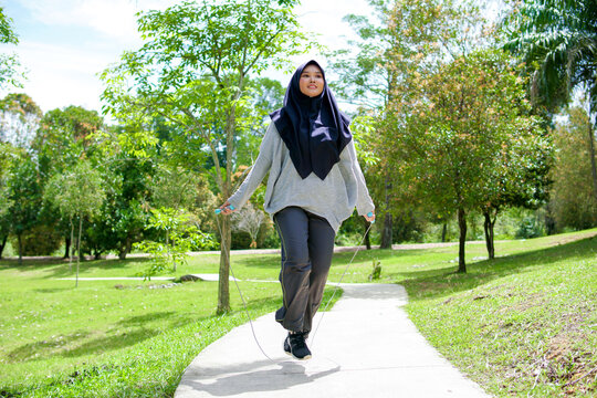 Picture of a young Muslim woman outside a green nature park doing exercises with a skipping rope.