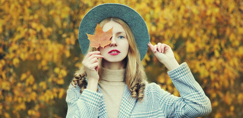 Autumn portrait of beautiful young woman covering her face with yellow maple leaves wearing a hat in park