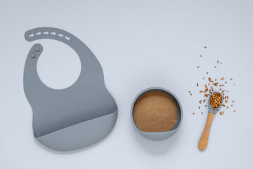 Flat lay composition of healthy baby food with a bowl, a spoon with a buckwheat and a bib or feeder on yellow background. The concept of baby care and first lure.