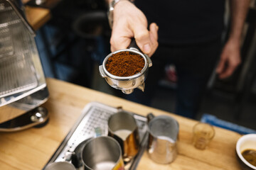 Bartender hand holding a portafilter with ground coffee on a blurred background with steel jugs