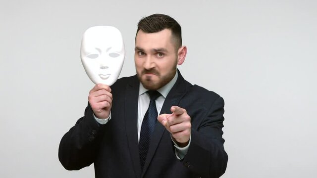 Bearded businessman in black official style suit pointing finger on you holding white mask with unknown face, blaming you in duplicity. Indoor studio shot isolated on gray background.