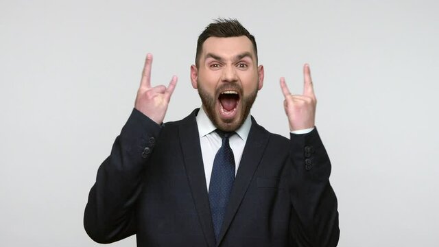 Portrait of overjoyed bearded businessman in black official style suit screaming loud with excitement and showing rock and roll gesture to camera. Indoor studio shot isolated on gray background.