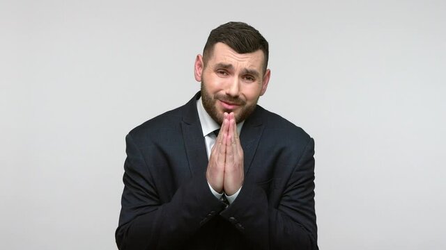 Please, I beg you! Hopefull bearded businessman in black official style suit praying for help, expressing big hope with imploring pleading eyes. Indoor studio shot isolated on gray background.