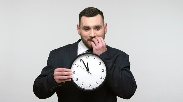 Nervous bearded businessman in black official style suit holding big wallclock and biting her nails, need more time for work, deadline. Indoor studio shot isolated on gray background.