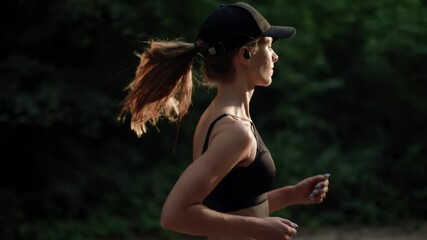 A young woman runner is listening to music in earphones and training in summer forest. Close up of fitness girl is jogging outdoor. Concept of workouts running and healthy lifestyle. Slow motion.