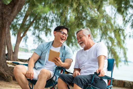 Happy Asian family handsome man teach senior father using digital tablet for video call or online shopping on the beach in summer day. Father and son enjoy and having fun together in holiday vacation