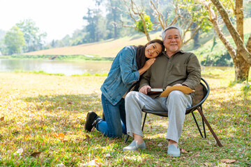 Asian woman caring and hugging senior man grandfather sitting on outdoor chair in the park. Elderly retired male relax and enjoy outdoor activity together with daughter. Family relationship concept