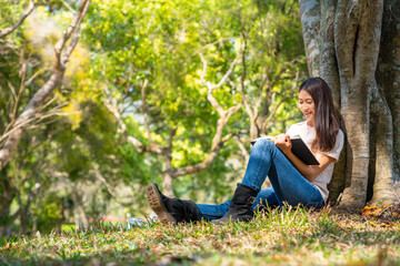 Young Asian woman sitting on grass under the tree reading a book in the park. Female college student relax and enjoy outdoor lifestyle activity in summer day. Education and leisure hobbies concept