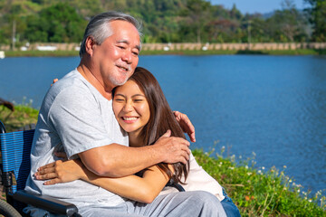 Happy Asian woman caring and hugging senior man grandfather sitting on wheelchair in the park. Elderly retired male relax and enjoy outdoor leisure activity with daughter. Family relationship concept.