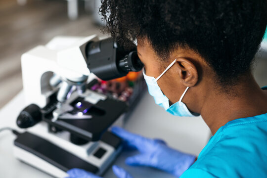Black medical technologist in mask in laboratory looking through microscope at blood samples