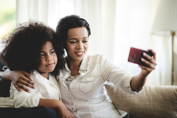 Cheerful young mother taking a selfie using smartphone with sister with curly hair while making funny facial and hand gestures sitting on couch at home