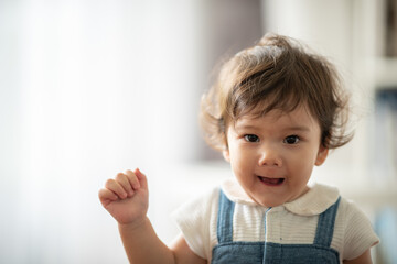 Cute little playful girl child learning to walk and stand by taking support of couch at home while looking away mischievously