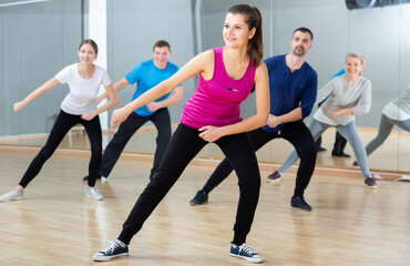 Fototapeta na wymiar Smiling young female coach doing dance workout with adult group in fitness center