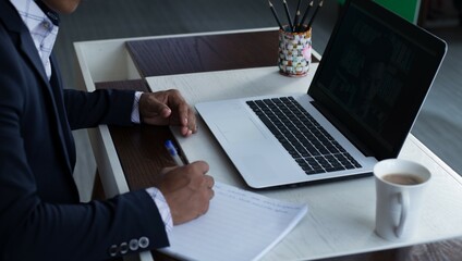 Young adult male writing down notes on a notebook. Unrecognizable businessman with laptop computer on the desk  working. 