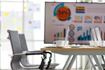 Laptop computers and white hot coffee cups on small dish placed on working table full of company paper report documents in meeting room in front of target growth rate graph and chart on big screen