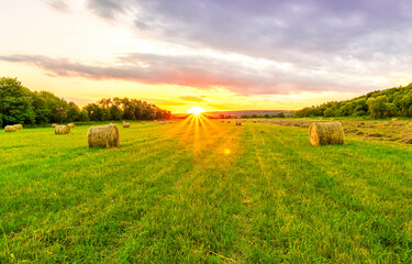 Obraz premium Scenic view at beautiful sunset in green shiny field with hay stacks, bright cloudy sky, country road and golden sun rays with glow, summer valley landscape