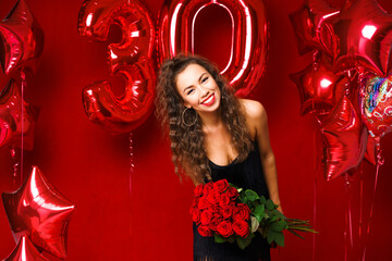 Obraz na płótnie Canvas Beautiful young woman on a red background with red balloons and a bouquet of red roses. Cheerful and happy girl with bright red lipstick celebrates 30 years anniversary