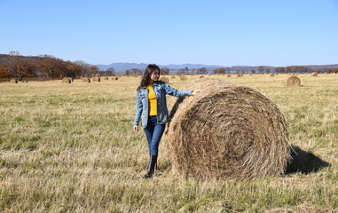 Young brunette woman in denim jacket standing near haystack in field with bales of hay