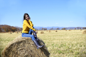 Young brunette woman in yellow sweater and rubber boots sitting on top of haystack in field