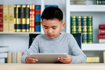 Asian boy on grey sweatshirt sit at school desk, lower head down to concentrate on online class study of kid lesson by reading ebook, info searching via tablet at library of elementary academy