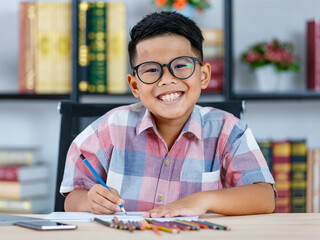 Lovely Asian boy wearing glasses and grey checked shirt sitting on desk of reading room and happily smiling while enjoy color drawing and art sketching on paper by blue pencil