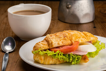 Breakfast consist of croissants with ham, cheese, slice tomato, onion and green vegetable in white ceramic dish and a cup of milk coffee.
