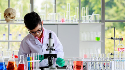 Asian schoolboy in glasses and lab coat examining chemical substance through microscope while conducting scientific experiment in school laboratory