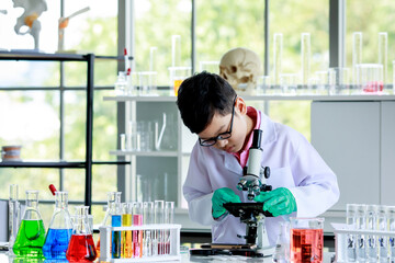 Concentrated smart Asian boy in lab coat and gloves with dropper pouring liquid sample under microscope lens while conducting experiment during chemistry research in school laboratory