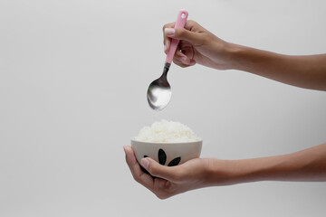 Hand holding a ceramic bowl of Cooked jasmine rice and spoon isolated on gray background.