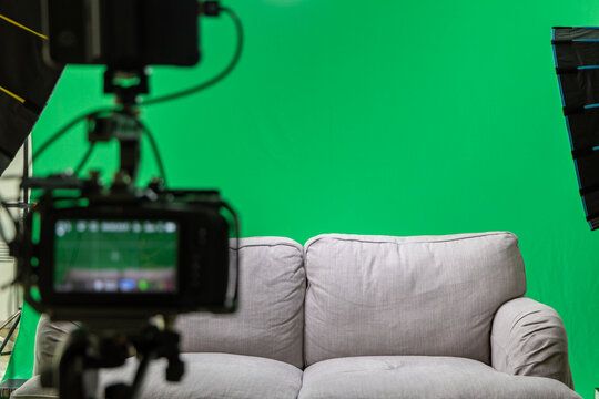 a gray couch stands in front of a green screen. Background interchangeable. in the foreground you can see a camera and softboxes. Interviewsetting with colored background. 