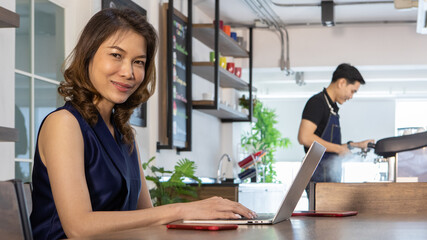 Pretty young Asian woman freelancer in dark tank top sitting near counter of barista in cafe on afternoon, turn to smile while typing on laptop as happy to work online business