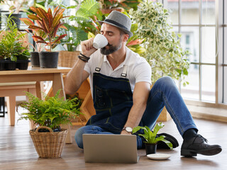 A bearded man wearing hat sitting and drinking coffee and using laptop notebook computer to sell...