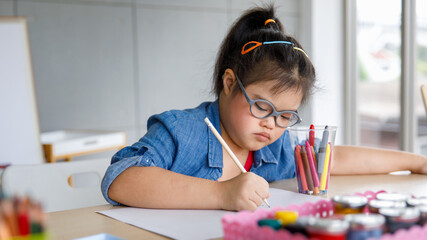 Young Asian down syndrome little girl sitting at desk with colors prepare for painting. She widely...