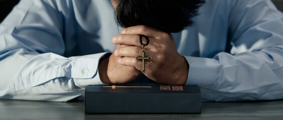 Asian man bowing his head on the Bible and carrying a cross. According to the beliefs of...