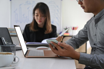 Businessman holding smart phone and working with laptop computer while sitting with his colleague in office.