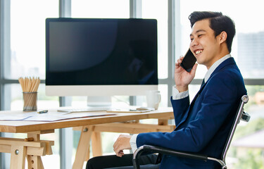 Asian young businessman sitting at desk and using smartphone to call and make communication near desktop computer