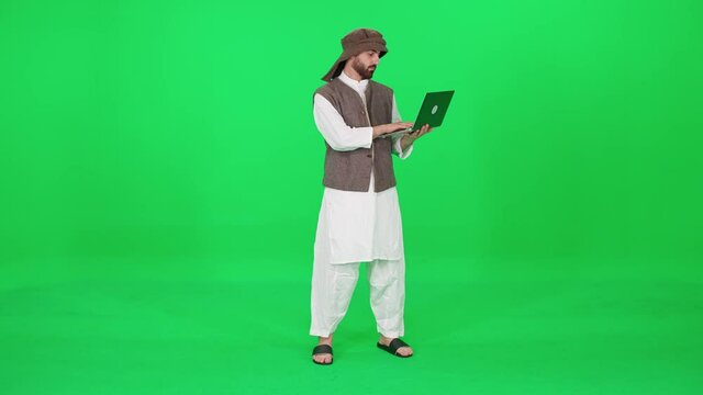 A man of Arab appearance in traditional clothes uses a laptop to surf the internet while stands against the background of a chromakey.