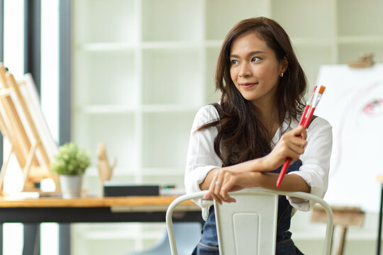 Pretty asian female artist sitting and holding painting brushes, smiling and day dreaming