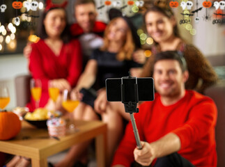 friendship, holiday and people concept - group of happy smiling friends in halloween costumes taking picture with smartphone on selfie stick at home party at night