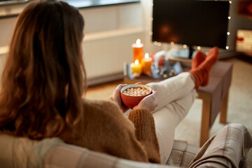 Fototapeta na wymiar halloween, holidays and leisure concept - young woman watching tv and drinking hot chocolate with marshmallow with her feet on table at cozy home