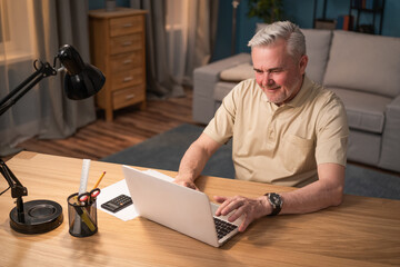 Fototapeta na wymiar Senior businessman holds online conferences. An elderly man works on his laptop in the evening at his desk and lamp. Mature businessman happy to work online. The man is working remotely.