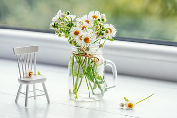 Wildflowers chamomile in a jar and miniature furniture on the windowsill